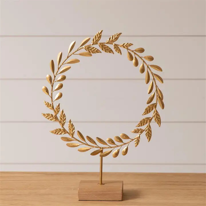 A flat-profile, gold leaf wreath mounted by a stem onto a light wood block.