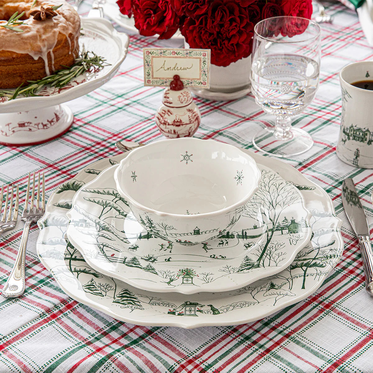 Christmas-themed table setting with white and evergreen toile dishes featuring a dinner plate, salad plate, and bowl on a red and green checkered tablecloth.
