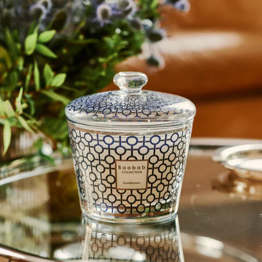 Decorative candle with glass lid and an intricate black design on a glass table with flowers in the background