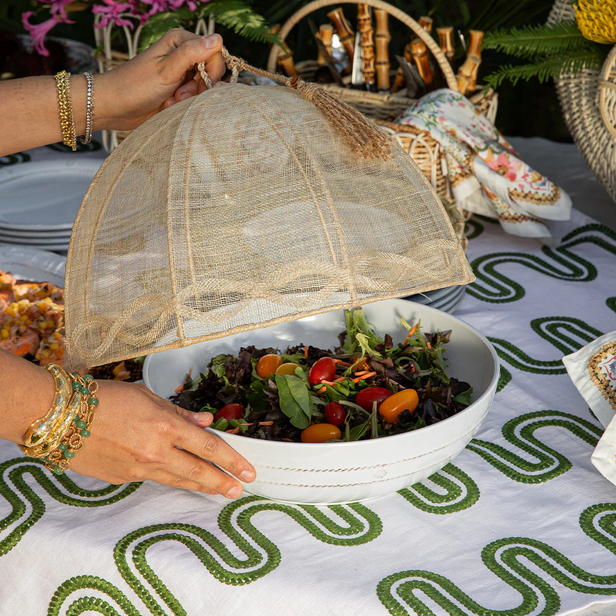 Person holding a mesh food cover over a bowl of salad on a patterned tablecloth.