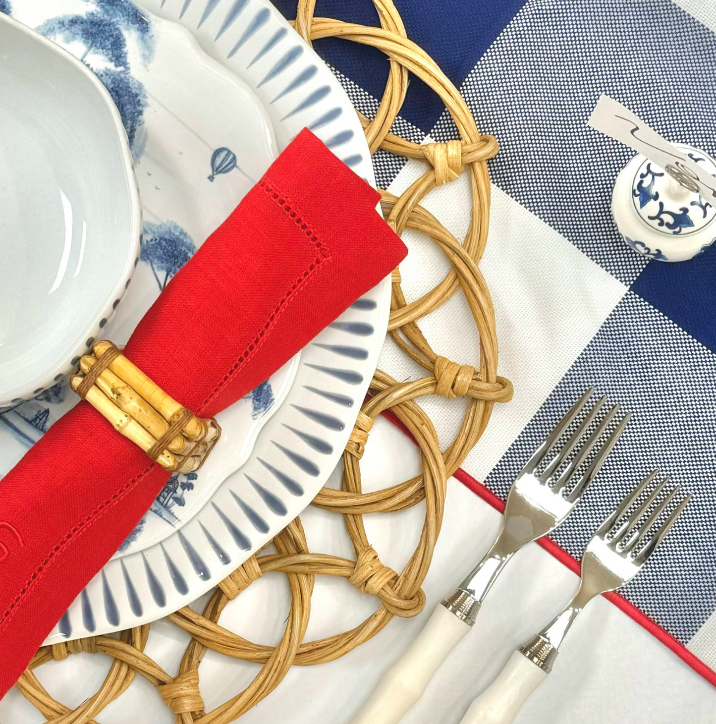 A rattan charger with an ornate, scalloped pattern poking out from under a red, white, and blue place setting on a blue gingham tablecloth
