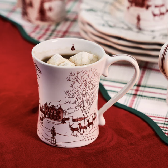White mug with red toile winter scene design filled with hot chocolate and marshmallows on a red tablecloth and other festive dishes