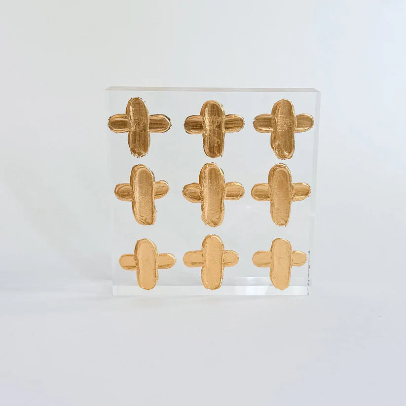 Nine abstract gold crosses on a clear acrylic block against a light gray background