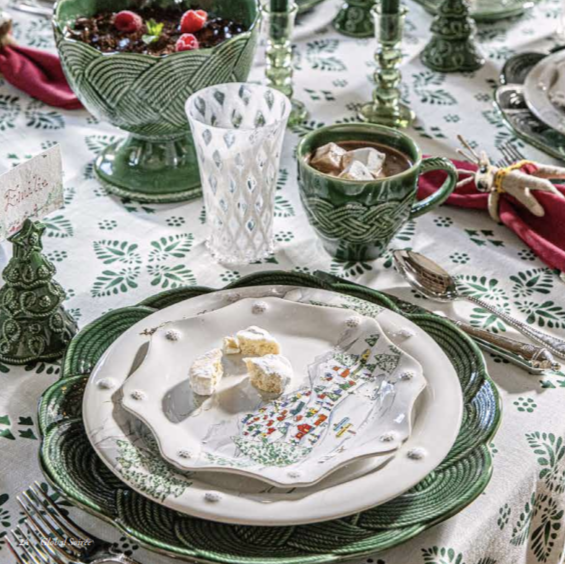 Festive holiday table setting with green and white dishes, red napkins, and a patterned tablecloth with varying Christmas motifs