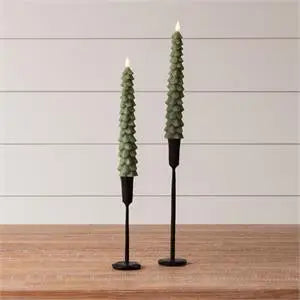 Two green tree-shaped candles in black candlestick holders on a wooden surface with a light gray background