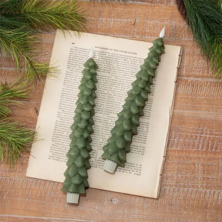 Two green Christmas tree-shaped candles with faux flames on an open book with pine branches around.