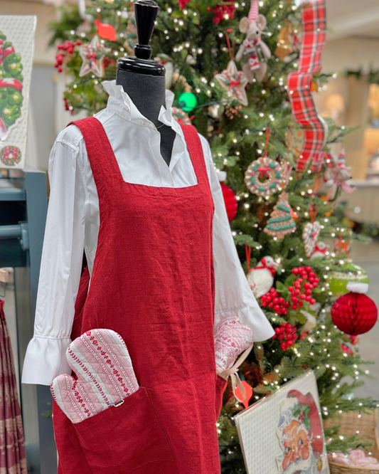 A bright red apron on a mannequin wearing a white button down standing in front of a festive Christmas tree holding red and white oven mitts.