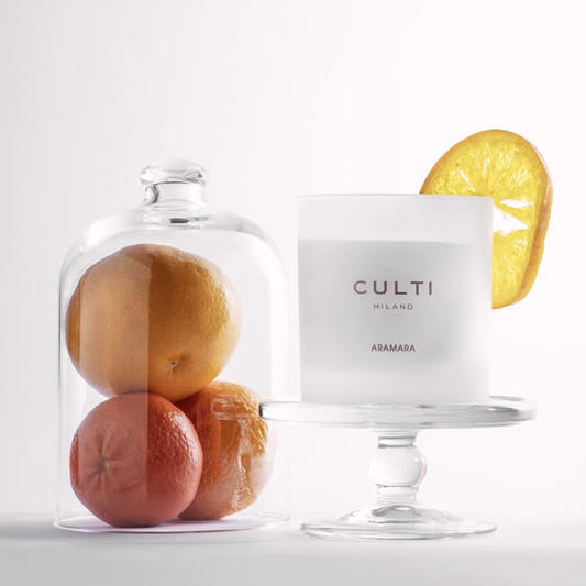 A crisp, white candle on a small cupcake stand next to a cloche filled with oranges on a white background