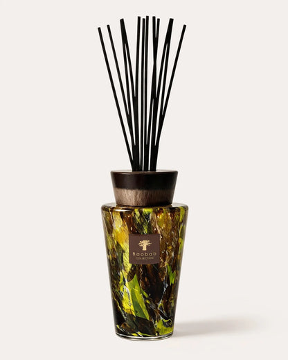 A reed diffuser bottle with a marbled green and yellow pattern, a dark brown wooden cap, and multiple black reeds, shown on a light beige background.