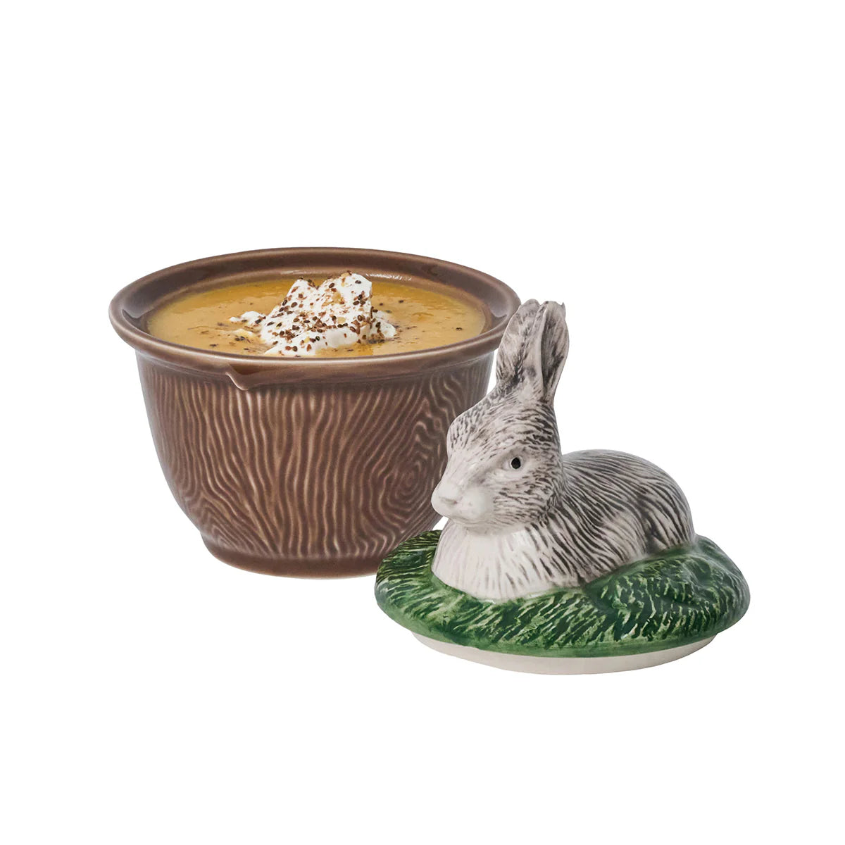 A brown-ceramic, faux bois bowl with soup positioned next to a lid with a rabbit laying in grass.