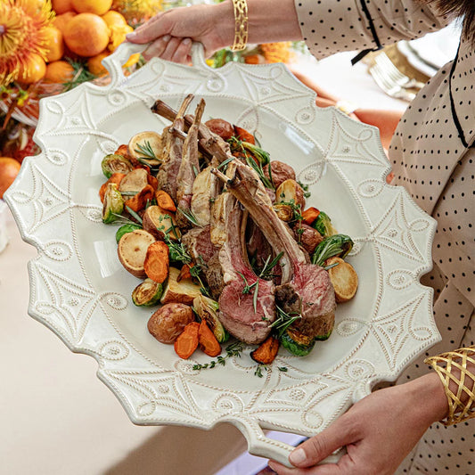 Decorative white platter with roasted lamb chops and vegetables held by a person.