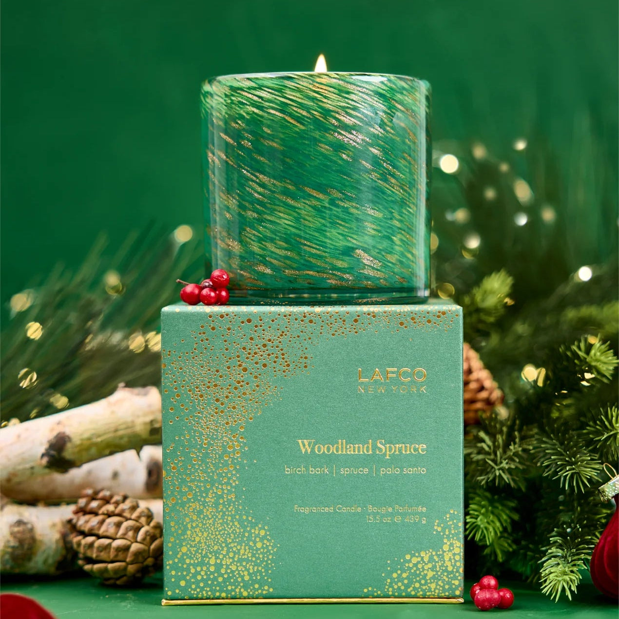 A green candle atop its corresponding green and bronze-foil box surrounded by small fire logs, pine garland, pinecones, and red ornaments.
