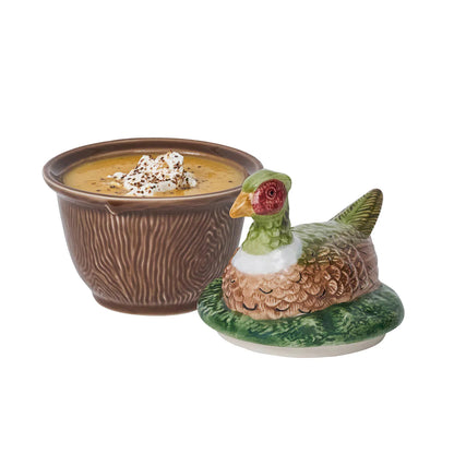 A brown-ceramic, faux bois bowl with soup positioned next to a lid with a pheasant laying in grass.