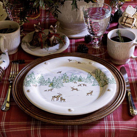Christmas-themed table setting with a festive plate featuring reindeer and trees on a dark wooden charger and red plaid tablecloth.