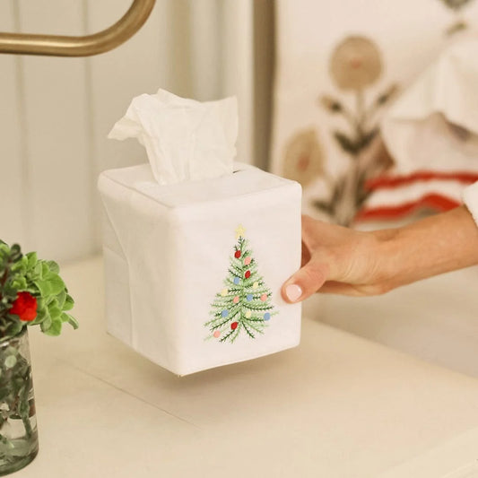A white square tissue box cover with an embroidered Christmas tree design, being held by a hand on a bathroom counter.